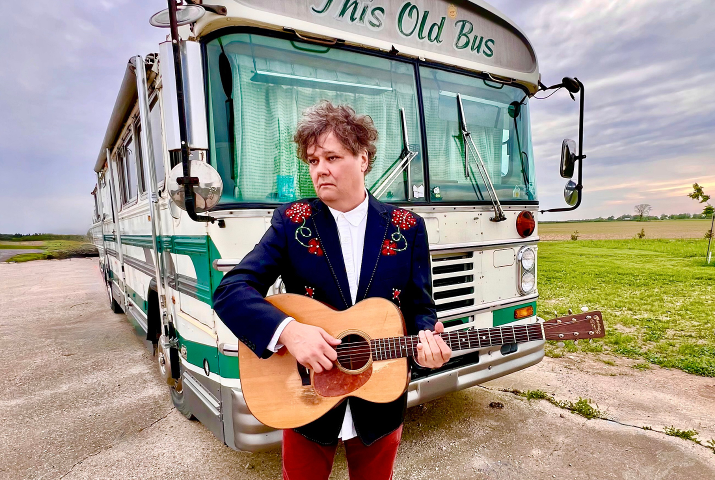 Artist Ron Sexsmith standing in front of old school bus with guitar